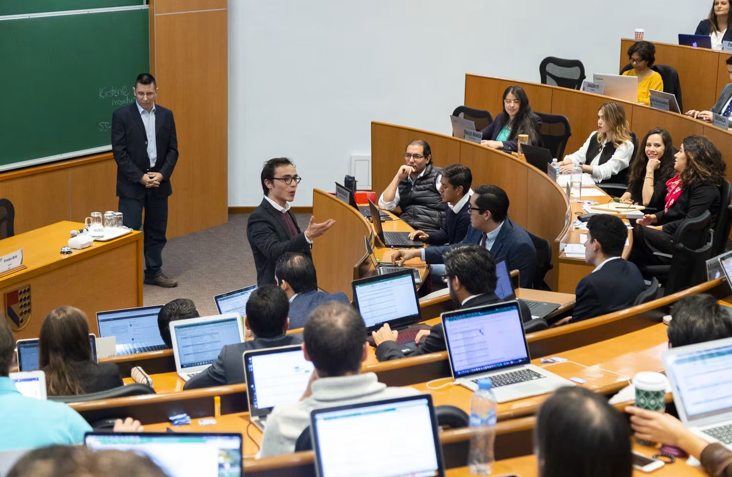 Sesión plenaria en aula de herradura: un participante de pie expone su postura mientras el catedrático escucha junto al pizarrón.